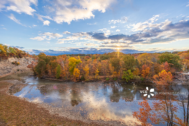 金色的阳光洒在宁静的湖面上，周围是色彩鲜艳的秋叶和白云蓝天。图片素材