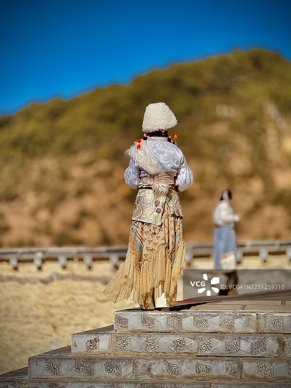 身着传统服饰的人站在台阶上，背景是山景和湛蓝的天空，这是从后面拍摄的。图片素材