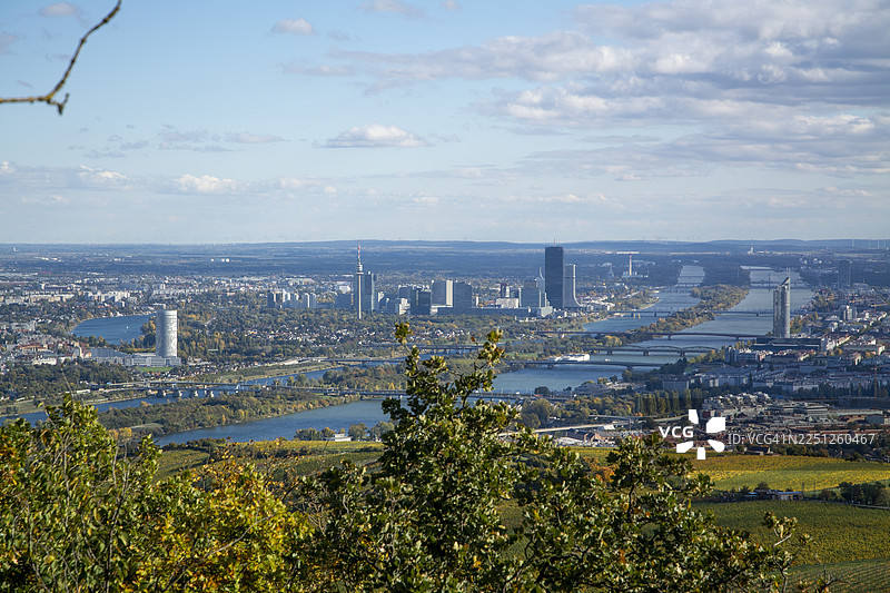 宽广的城市天际线航拍，现代建筑沿宽阔的河流延伸，周围环绕着郁郁葱葱的绿色和秋色树木，天空部分多云，地点：维也纳，奥地利，多瑙岛图片素材