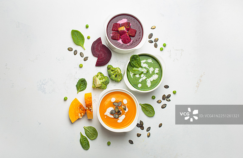 浅色背景上三碗色彩鲜艳的蔬菜汤，配有新鲜食材，俯视拍摄。图片素材