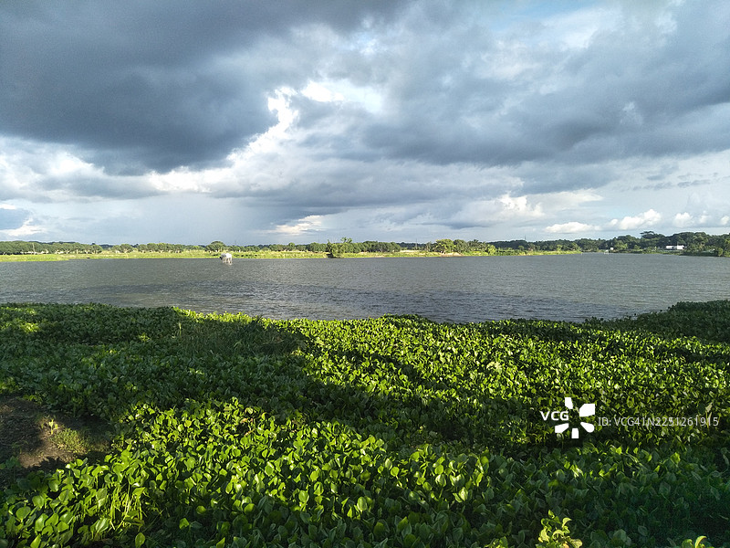 宁静的湖泊被青翠的植被覆盖，远处的地平线勾勒出树木的轮廓，头顶是戏剧性的云彩天空。图片素材