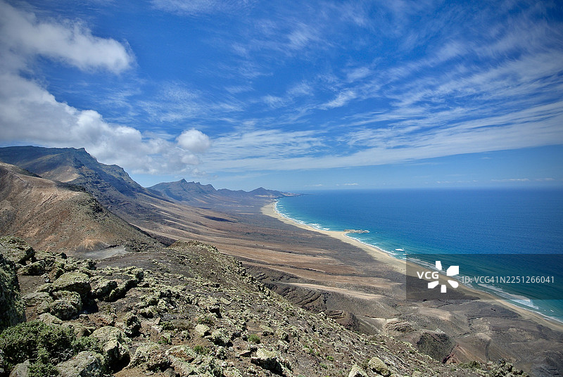 西班牙富埃特文图拉岛，崎岖的山脉与原始海滩和广阔的蓝色大西洋在充满活力的天空下交汇，构成壮丽的海滨风光图片素材