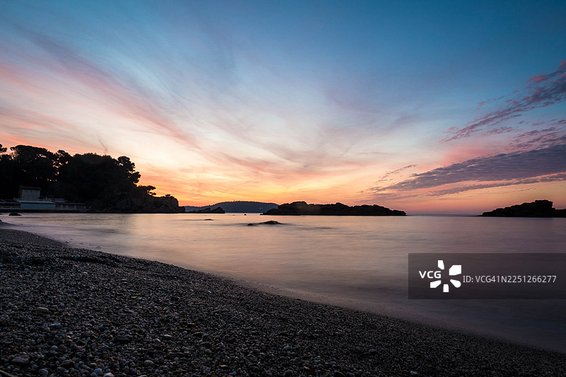法国土伦，宁静的日落映照在鹅卵石海滩上，海面平静，远方是剪影般的海岸线图片素材