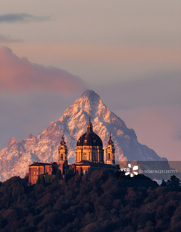 夕阳映照下，雄伟的苏佩尔加大教堂坐落于森林覆盖的山丘上，背景是白雪皑皑的山峰和粉红色的云彩，意大利，圣拉斐尔·西梅纳图片素材