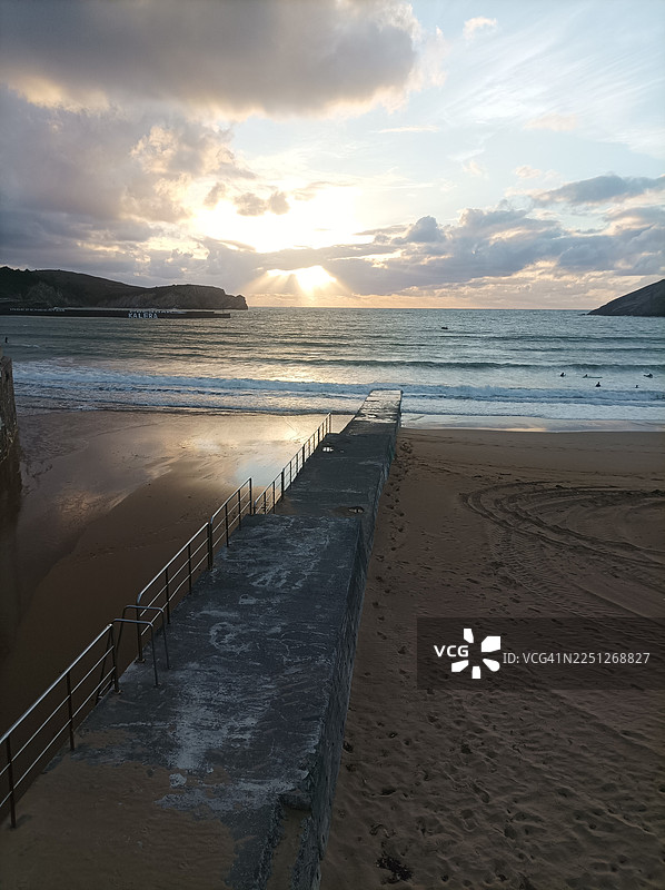 金色日落光芒照亮宁静的海滩，海边的码头延伸至轻柔的海浪和远处的山丘，西班牙比斯开省图片素材