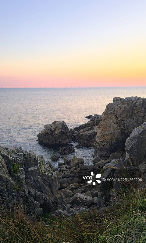 法国勒克鲁瓦西，日落时分，清澈天空下的岩石海岸风景，远方地平线上有风力涡轮机图片素材