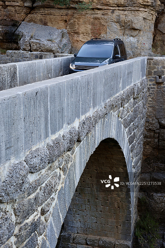 一辆深灰色的汽车正在穿越一座古老的石头拱桥，周围环绕着崎岖的自然岩石构造。图片素材