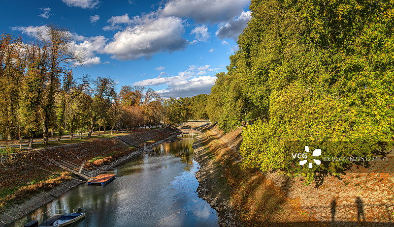 秋日下，一条宁静的河流蜿蜒穿过树林，明净的蓝天下点缀着蓬松的云朵，构成一幅绝美的风景画。图片素材