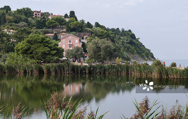 斯洛文尼亚菲耶萨，清澈天空下，绿树覆盖的山坡上坐落着一座迷人的粉色建筑，湖面倒映出这迷人的风景。图片素材