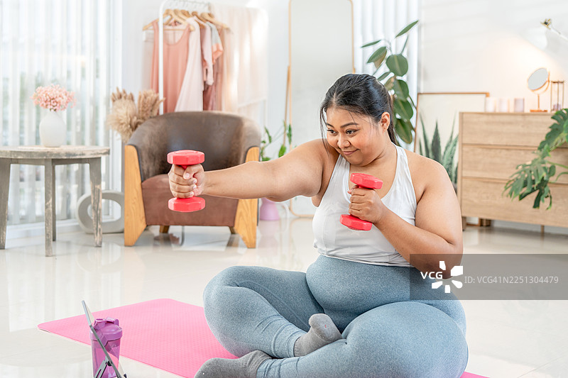 活动锻炼健康的身体，用于饮食或肥胖的概念。一个快乐的超重胖女人手里拿着哑铃，在房间里锻炼，进行饮食减肥。图片素材
