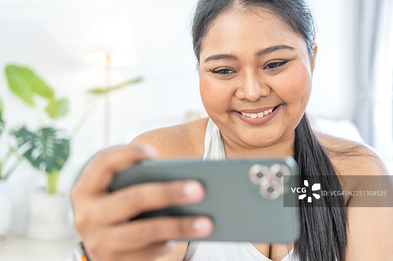 年轻的亚洲肥胖健身女性在健身房使用智能手机放松。超重女孩在锻炼后休息。肥胖女性在休息。胖乎乎的放弃。生活方式图片素材