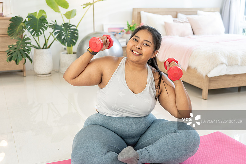 健身活动，为减肥或肥胖概念打造健美体魄。一位快乐的超重肥胖女性，双手持哑铃，在房间里锻炼，进行饮食和减肥。图片素材