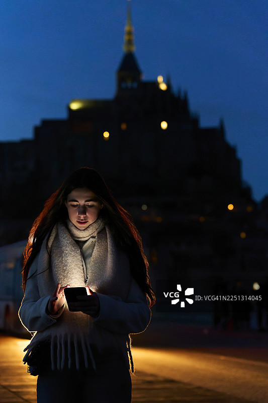 夜色中，年轻女子在圣米歇尔山前使用智能手机图片素材