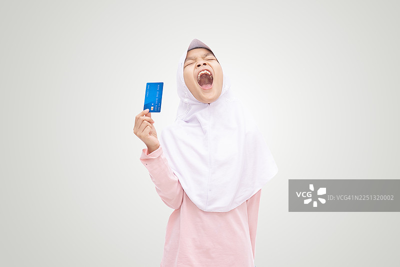 一个快乐的年轻穆斯林女孩，戴着头巾，手里拿着信用卡，在白色背景上兴奋地微笑，代表着财务自由、储蓄、教育、家庭财务和现代伊斯兰生活方式。图片素材