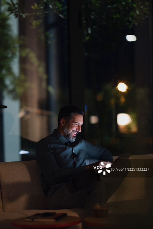 夜间，一位男士在现代家居环境中，舒适地躺在沙发上，使用平板电脑。图片素材