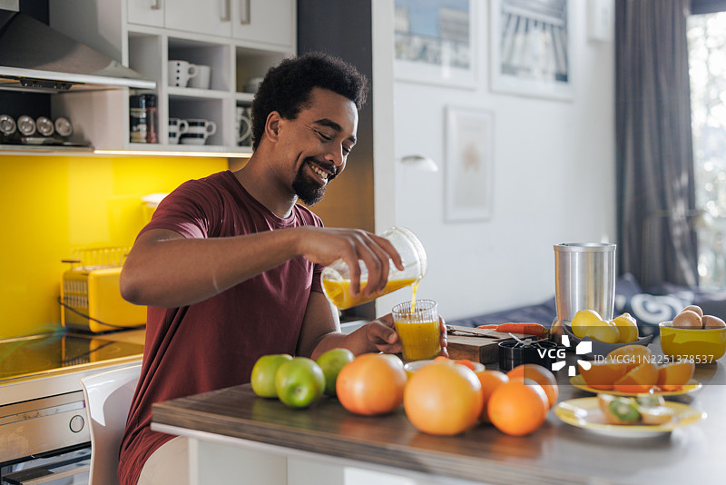 年轻男子在现代家居厨房里将新鲜果汁倒入玻璃杯中，台面上摆放着水果图片素材