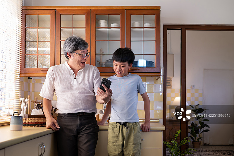 快乐的亚洲爷爷和孙子在厨房里使用智能手机图片素材