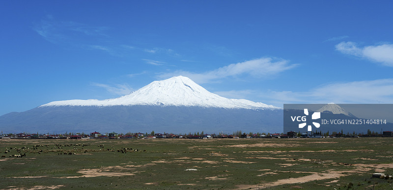 被绿色田野环绕的小村庄上方是雪山，天空湛蓝，远眺阿拉拉特山，包括大阿拉拉特山、小阿拉拉特山，位于土耳其东安纳托利亚地区的道乌拜亚泽特（阿勒省）。图片素材