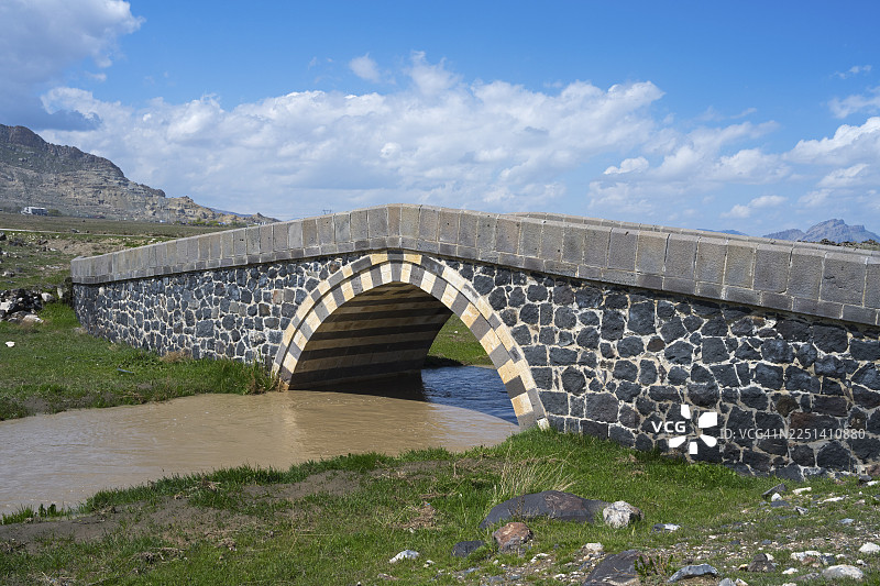 土耳其东部安纳托利亚地区阿勒省（Ağrı）的 Doğubayazıt（切夫尔梅附近）的石桥，坐落于蓝天下的绿色景观河流之上图片素材