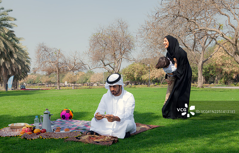 阿拉伯家庭在绿色公园野餐，父亲使用智能手机，母亲和孩子在一旁玩耍图片素材