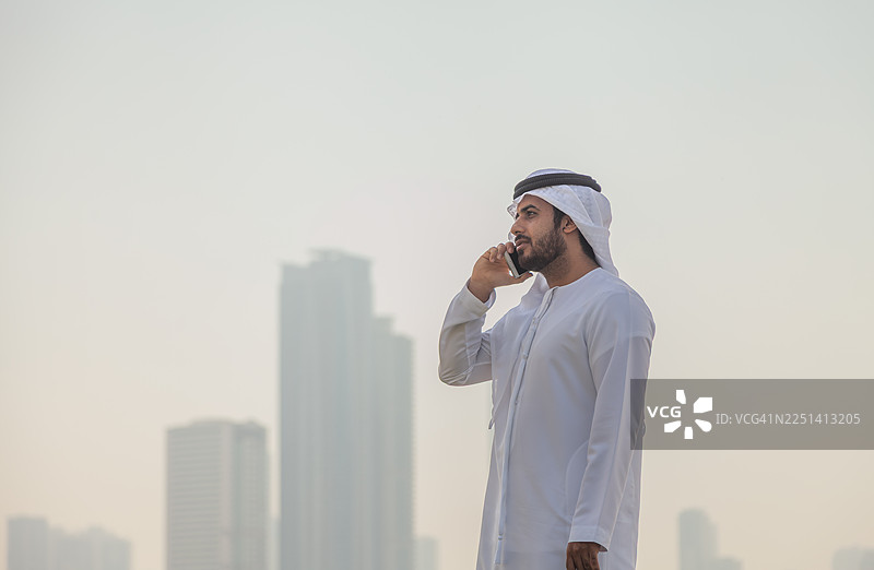 传统阿联酋男子在滨水公园用手机通话，背景是迪拜天际线图片素材