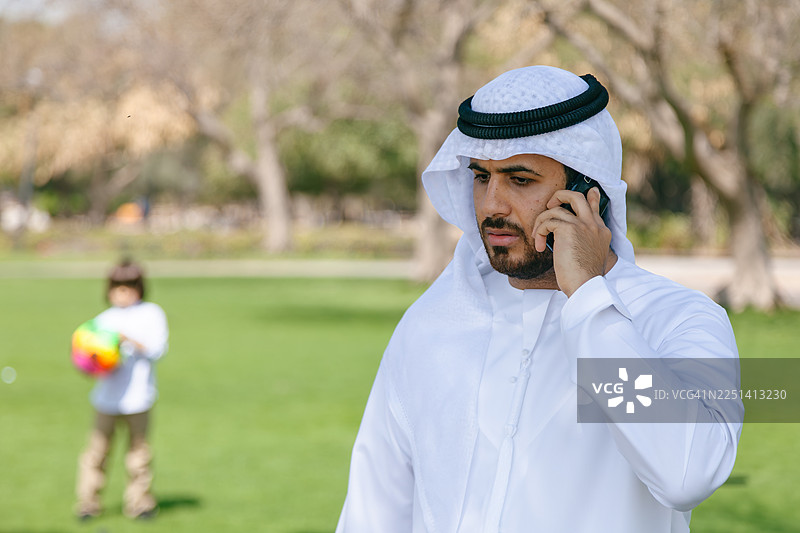 阿拉伯中东父亲在迪拜阿联酋的公园里，一边玩手机一边监督孩子玩球图片素材