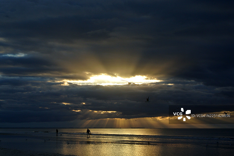 海边戏剧性的日落，阳光穿透乌云，海滩上有剪影人物图片素材