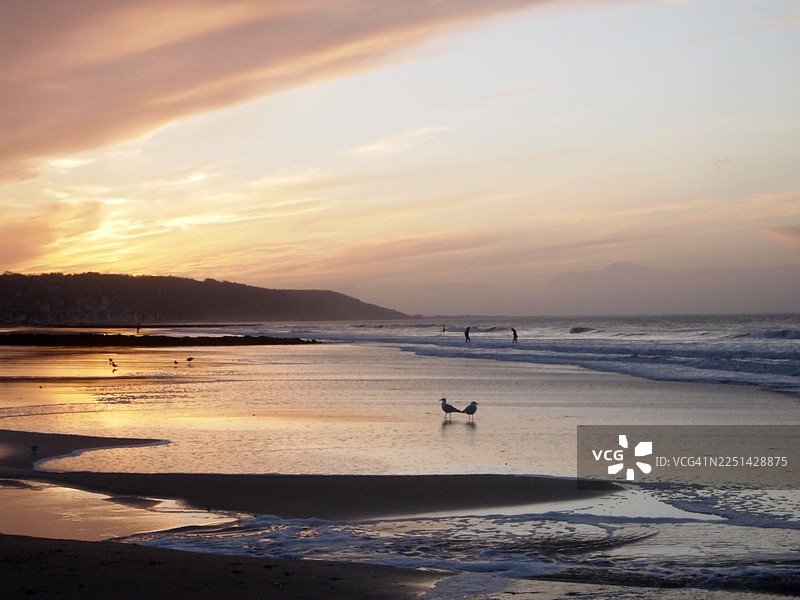 法国宁静的日落，广阔的海滩，轻柔的海浪和远方的人物，温暖的色调倒映在湿润的沙滩上图片素材