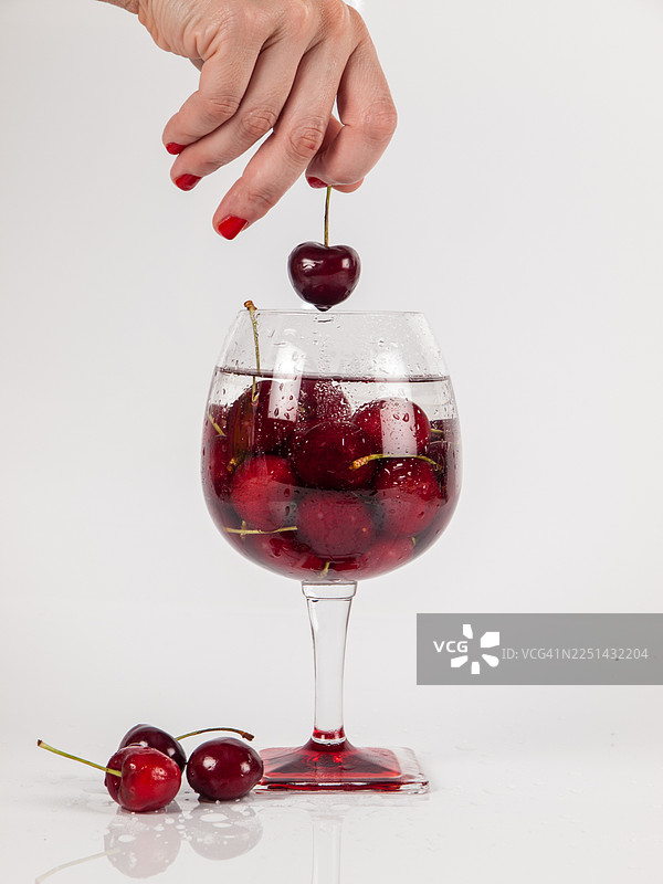 一只手将一颗新鲜的红樱桃放入装满樱桃和水的玻璃杯中，背景是干净的白色图片素材