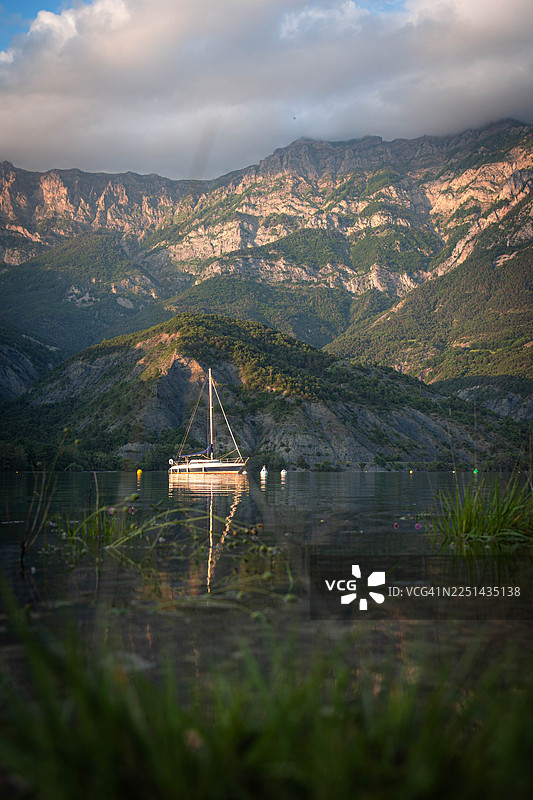 法国科尔日，日落时分，宁静湖面上帆船的风景，背景是雄伟的山脉和多云的天空图片素材