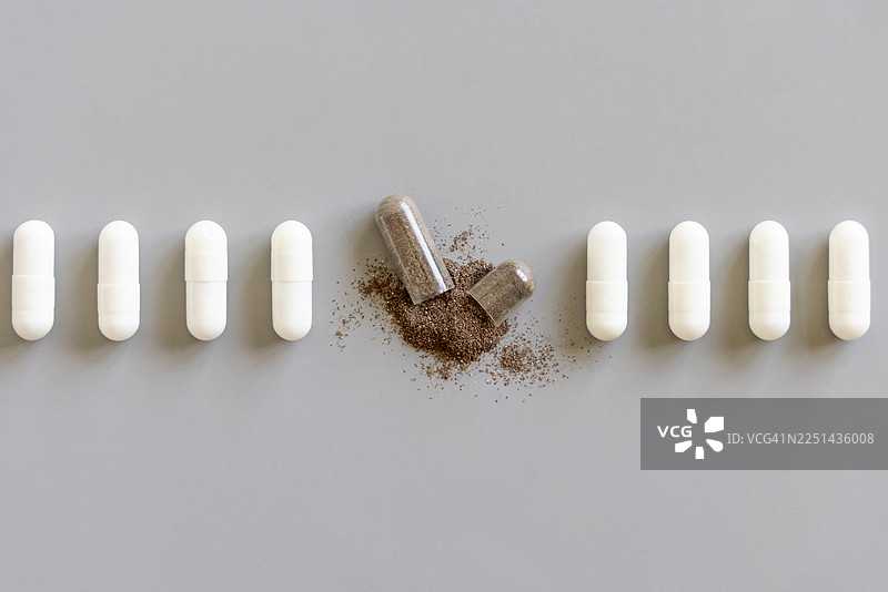灰色背景上，一排白色胶囊，其中一个破裂，内容物洒出图片素材