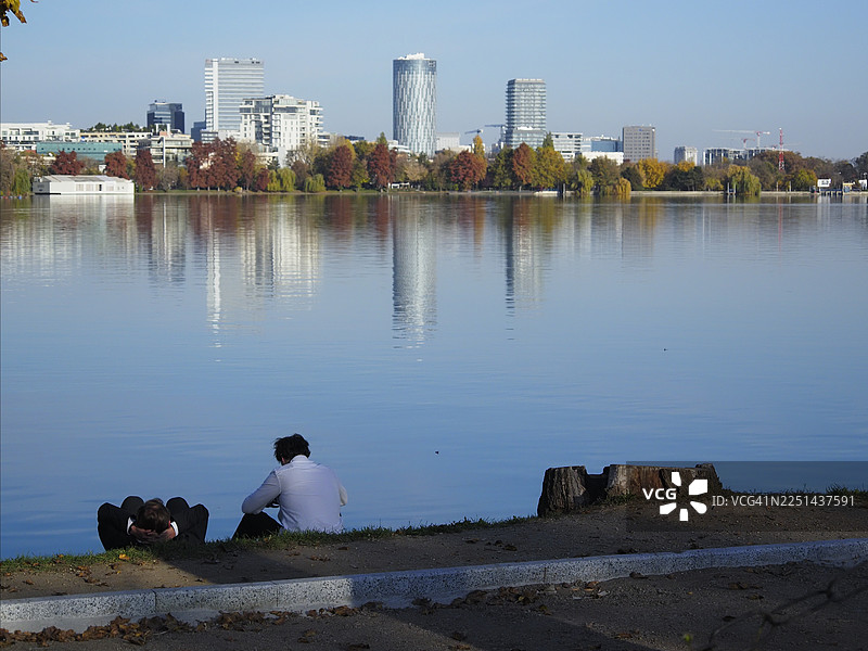 晴朗的一天，两个人放松地在宁静的湖边，背景是城市天际线，罗马尼亚布加勒斯特图片素材