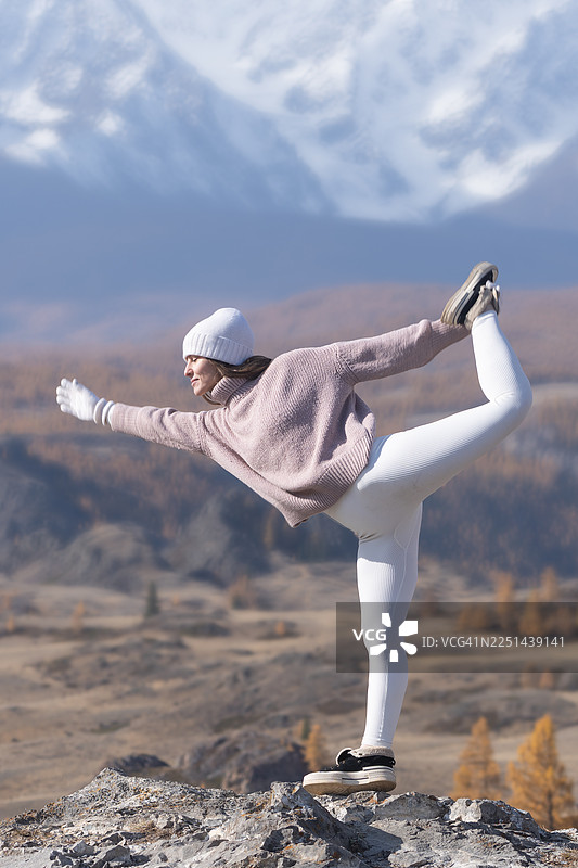 一位女子在山顶练习瑜伽，在白雪皑皑的山峰背景下保持着瑜伽体式平衡。图片素材