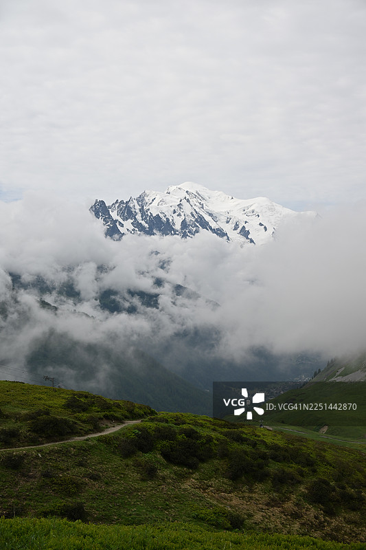 法国博纳维尔，被云雾笼罩的雄伟雪山，前景是穿过绿色山丘的蜿蜒小路图片素材
