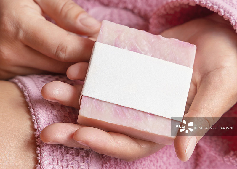 粉色毛巾背景下，一位女士手中拿着一块贴有空白标签的粉色香皂的特写。图片素材