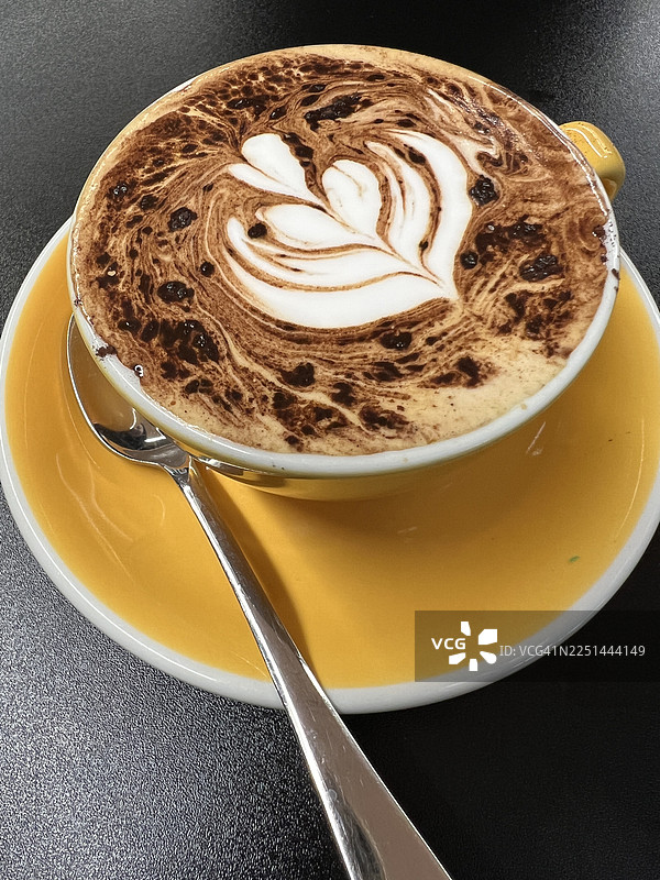 俯视角度拍摄的精美卡布奇诺咖啡，带有拿铁艺术，盛放在黄色杯碟中，置于黑色桌面上图片素材