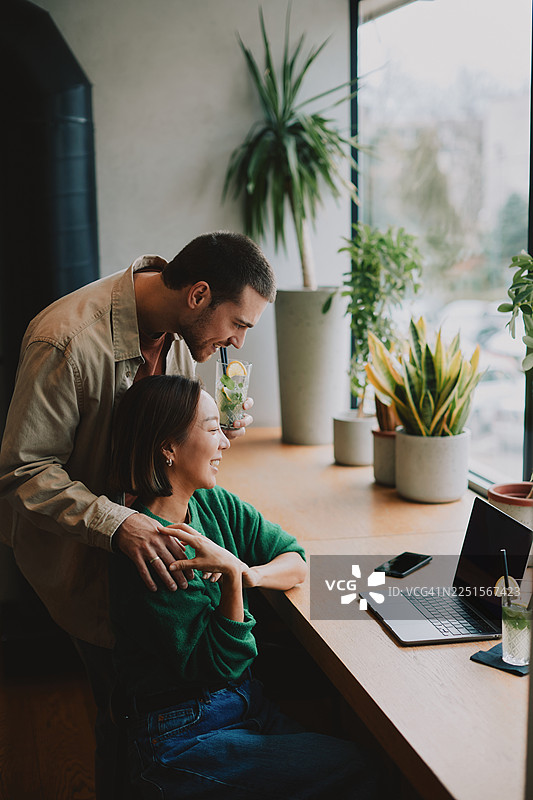 一对幸福的年轻已婚夫妇在种满植物的舒适咖啡馆里享用饮品并使用笔记本电脑图片素材