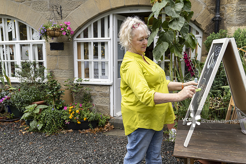 一位年长的女艺术家正在用粉笔为她的艺术工作室创作一个“Open”标志。图片素材