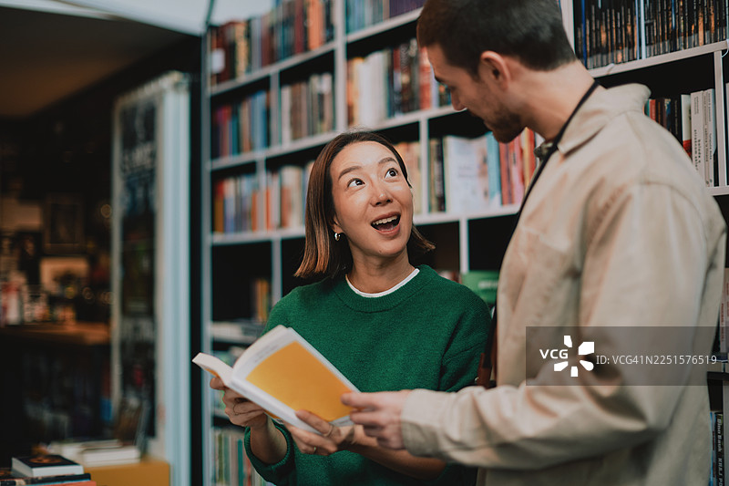 两位成年人在图书馆五颜六色的书架间一同阅读一本书图片素材