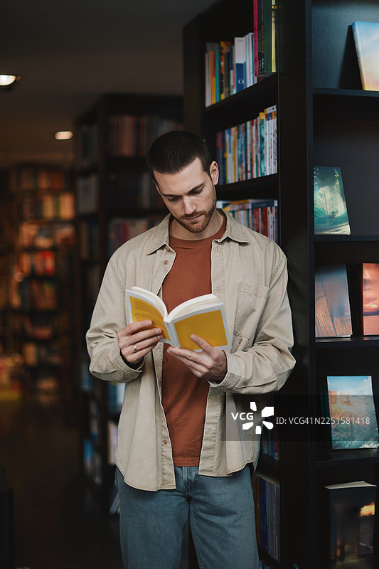 一位快乐的成年男子在舒适的图书馆里阅读一本黄色的书，面带微笑，享受悠闲时光图片素材