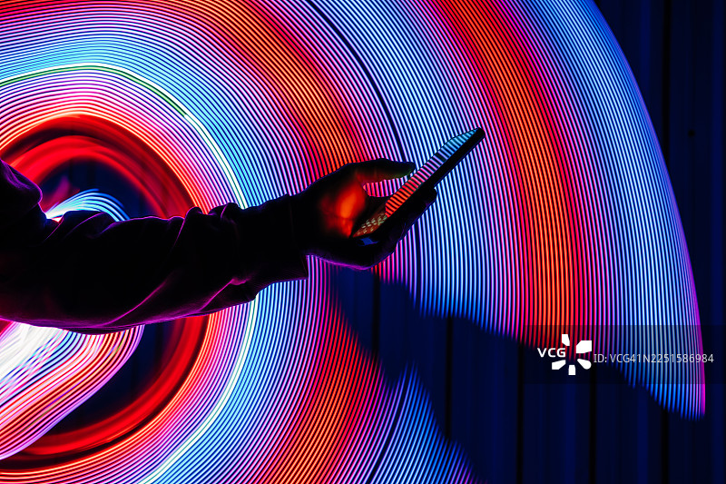 一只手举着智能手机，背景是动态的霓虹灯光轨迹。充满活力的长曝光效果象征着高速连接、数字创新和现代科技。图片素材