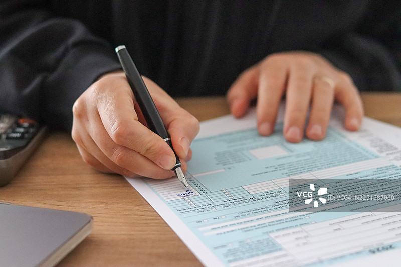 在家庭办公空间中，特写镜头展示了双手正在整理税务表格和财务文件，旁边有计算器、笔记本电脑和钢笔。自然光照亮了用于商业和编辑目的的税务准备和个人财务管理的详细过程。图片素材