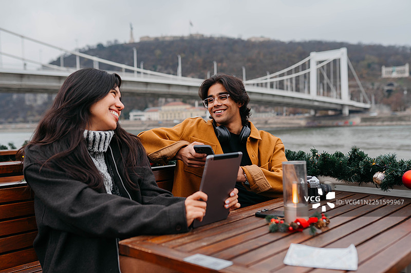 一对情侣在布达佩斯河畔的户外咖啡馆享受冬日午后，他们一边使用平板电脑，一边分享着笑容。图片素材