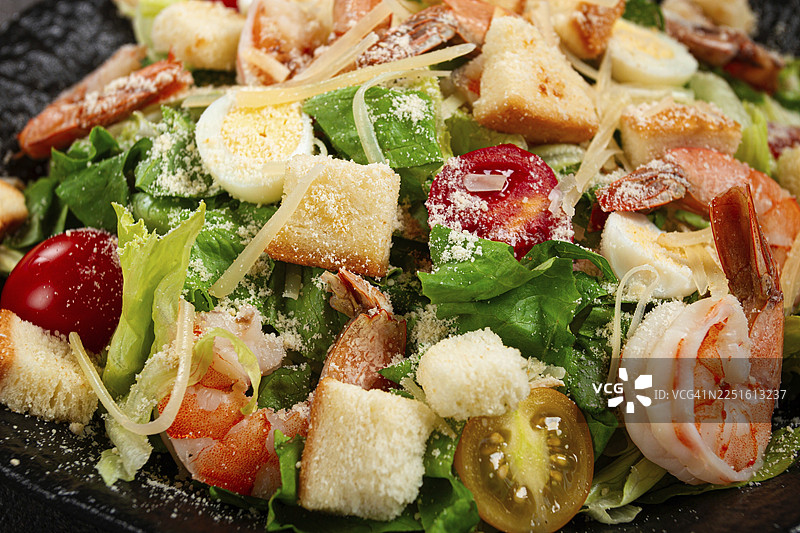 虎虾凯撒沙拉，包含虾、生菜叶、鹌鹑蛋、圣女果、帕玛森芝士，盛放在黑色盘子里图片素材