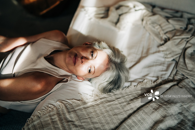 一位白发中年女性躺在床上，明亮的阳光洒在她脸上。主题为早晨的沉思、优雅地老去以及独处的宁静之美。图片素材