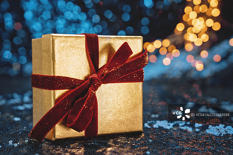 节日蓝色背景上带有金色灯光和闪光散景效果的优雅金色礼品盒，系有红色丝绒蝴蝶结，可用于企业礼品或奖励的摹仿。图片素材