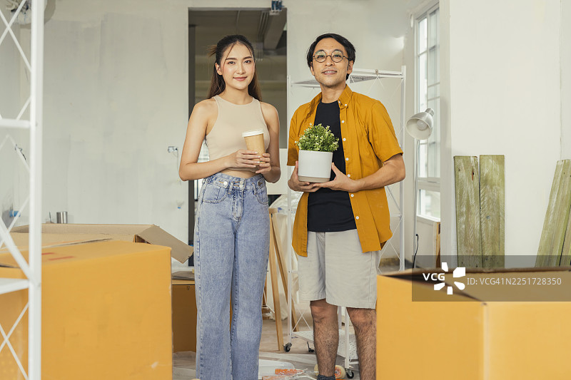 一对年轻的亚洲情侣穿着休闲服，在新公寓里整理和拆箱组装家具，进行DIY翻新和搬家，开心地展示着箱子并憧憬着他们的未来家园。图片素材