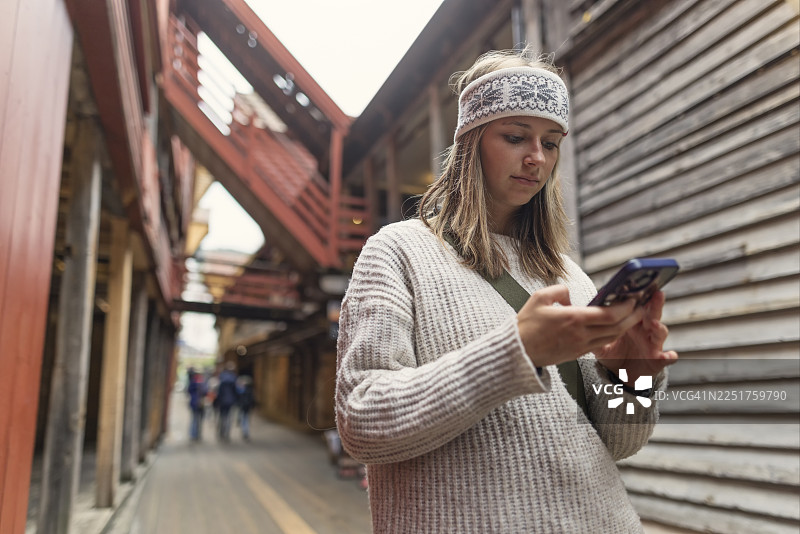 年轻女子在卑尔根探索布吕根的木制小巷，询问她的人工智能助手有关该区的历史图片素材