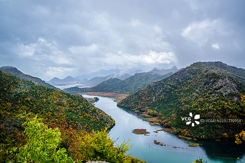 黑山斯卡达湖的秋季全景，宁静的河道蜿蜒穿过森林覆盖的山丘，色彩斑斓的树叶、芦苇岛、远处的山脉以及天空中飘浮的柔和云彩。图片素材