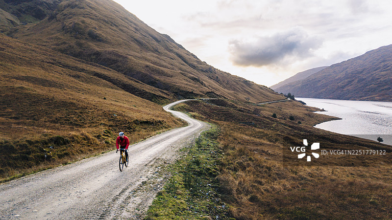 成熟男子在苏格兰高地湖畔的砂石小径上骑行的航拍/无人机视角图片素材